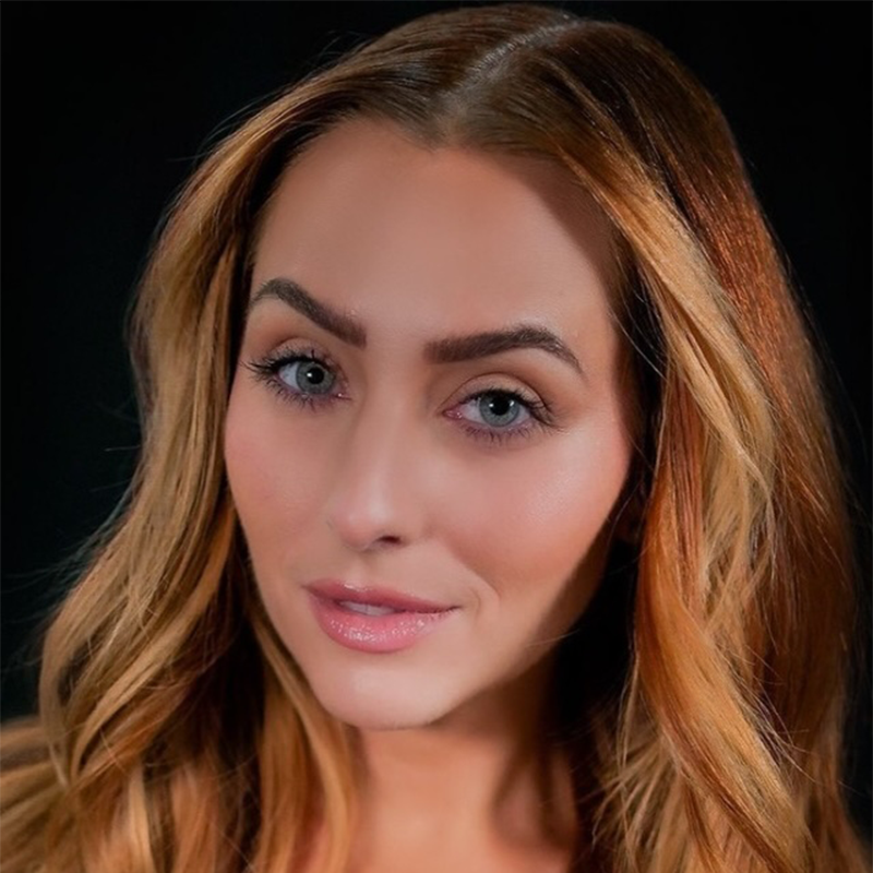 Kelsie Watts, a woman with long, wavy, light brown hair and fair skin, faces forward against a black background. She has blue eyes, defined eyebrows, and wears natural makeup with pink lipstick. The lighting highlights her features.