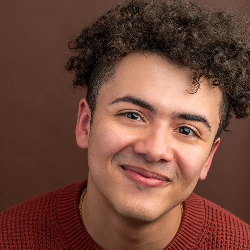 A young man with curly brown hair smiles gently at the camera. He is wearing a rust-colored knit sweater and a gold chain. The background is a plain, warm brown, and the lighting highlights his clear skin and expressive eyes.