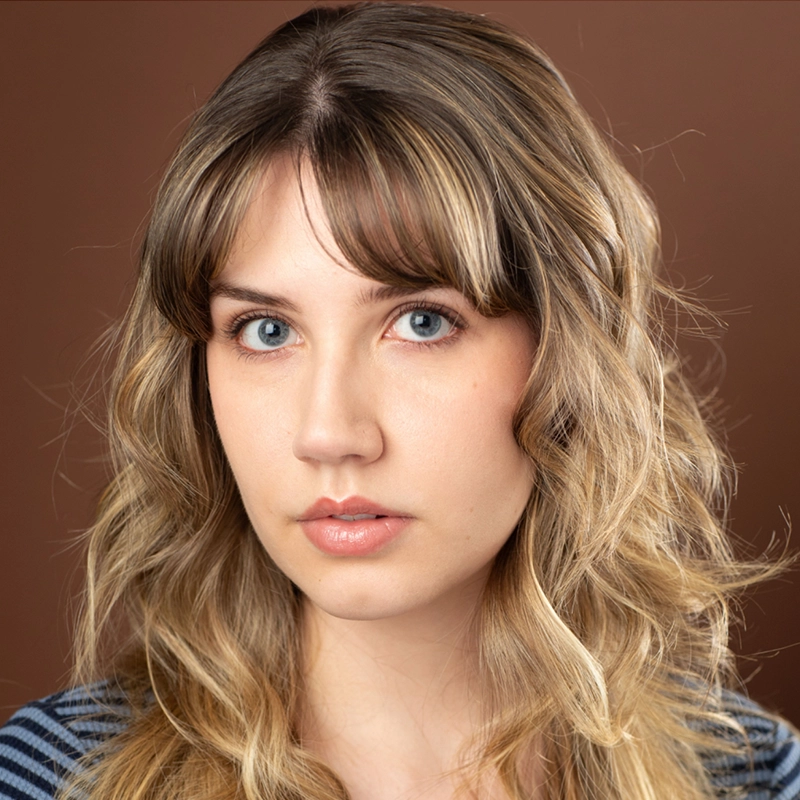 A young woman with wavy, shoulder-length blonde hair and bangs poses against a brown background. She has light skin, blue eyes, and wears a blue and white striped top. The lighting is soft and even—she resembles Taylor Leccese.