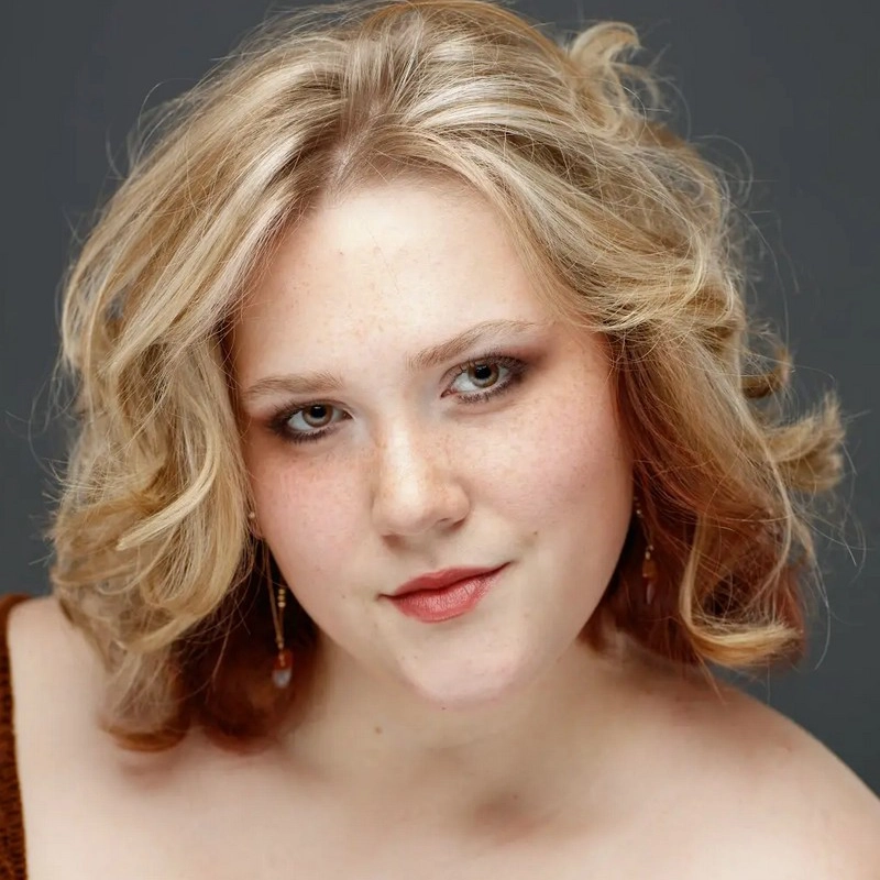 A young woman with light skin, wavy blonde hair, and brown eyes looks at the camera with a gentle smile. She is wearing subtle makeup, dangle earrings, and a brown off-shoulder top. The background is a plain dark gray. Deryn Kraner.
