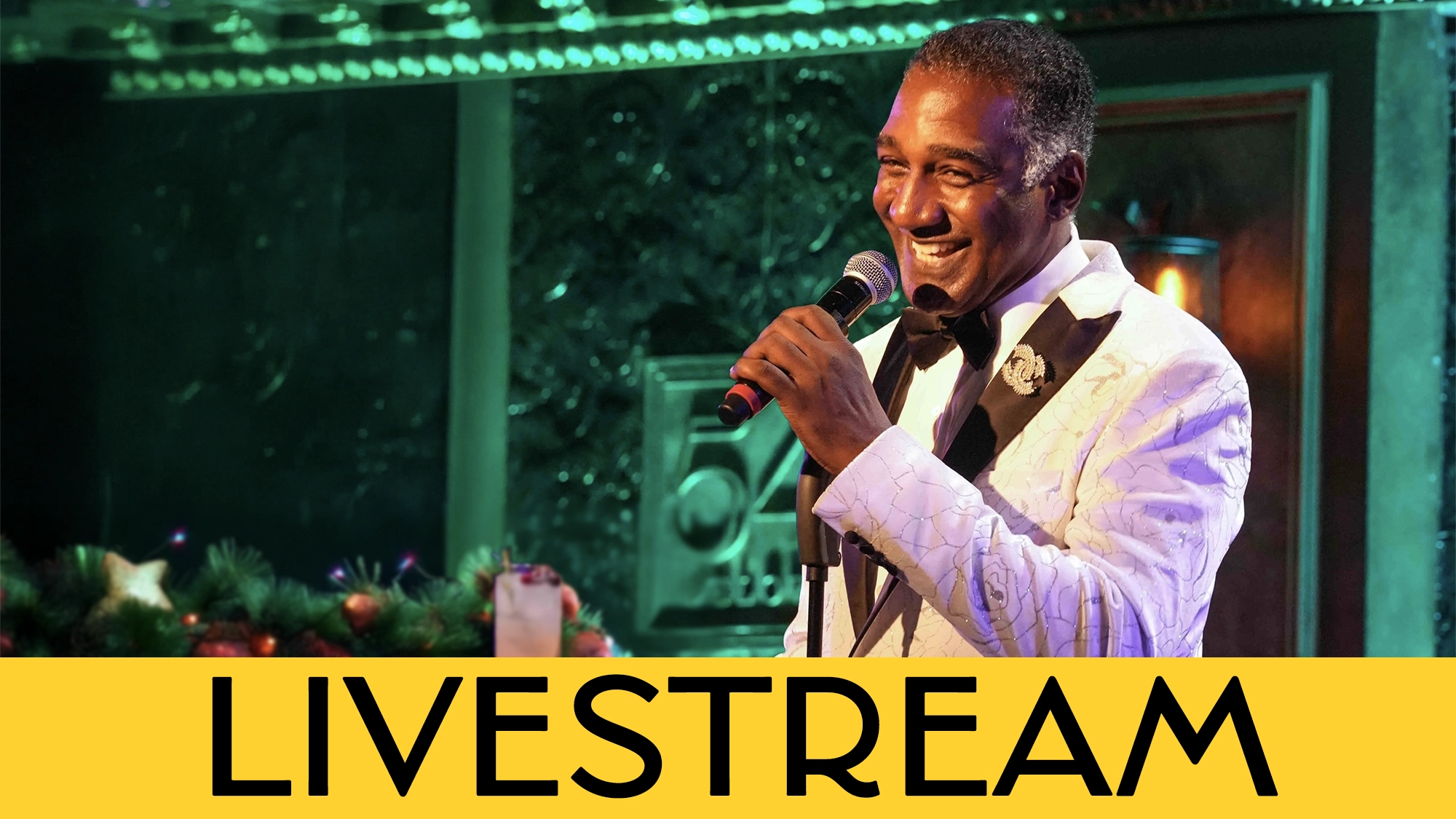 A smiling Norm Lewis in a white tuxedo jacket and black bow tie sings into a microphone on a greenery-adorned stage. The word 