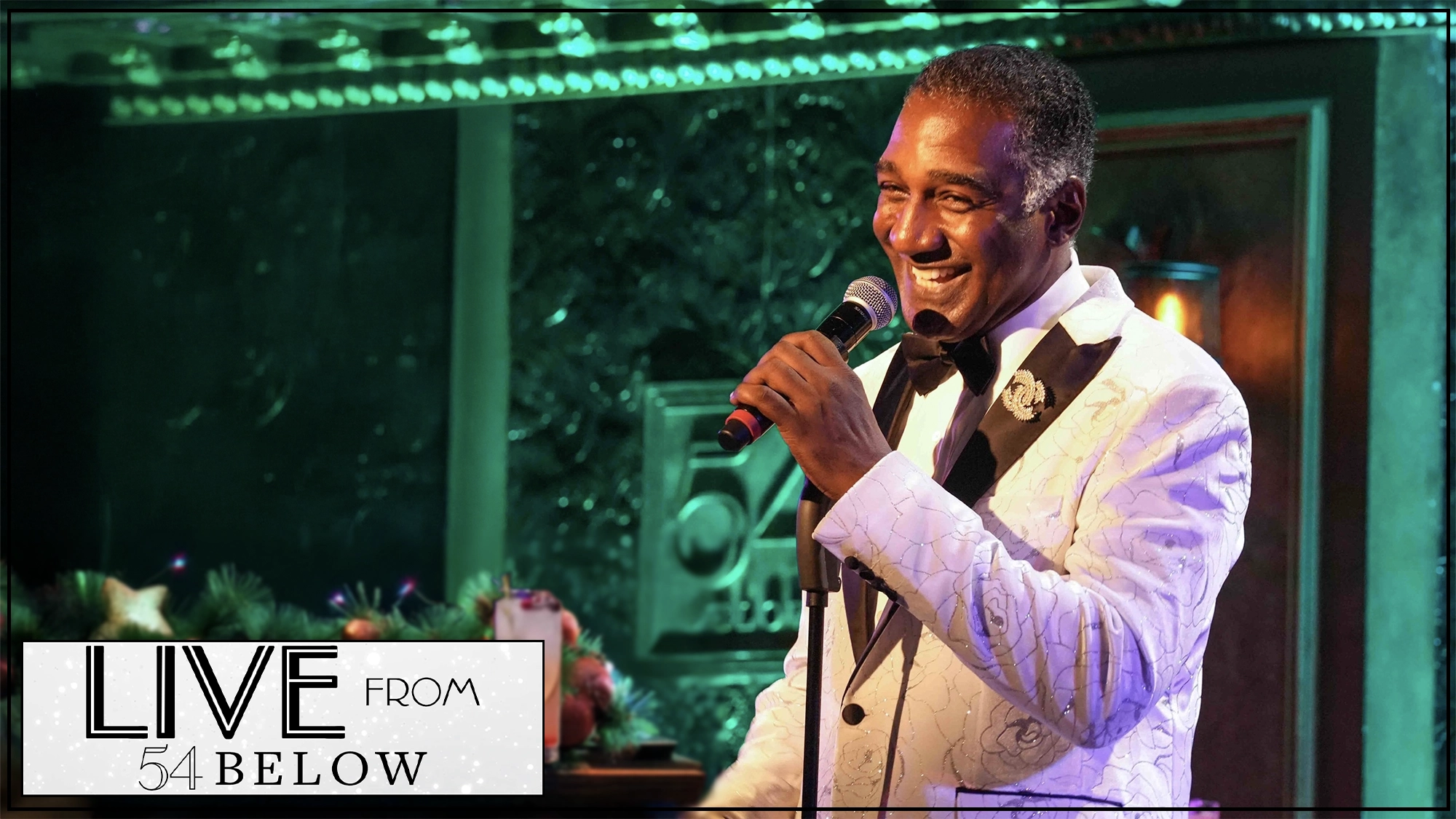 A smiling Norm Lewis in a white tuxedo jacket holds a microphone, performing on stage at 54 Below. The warmly lit background glows green and pink, capturing the holiday spirit of his Christmas livestream. 