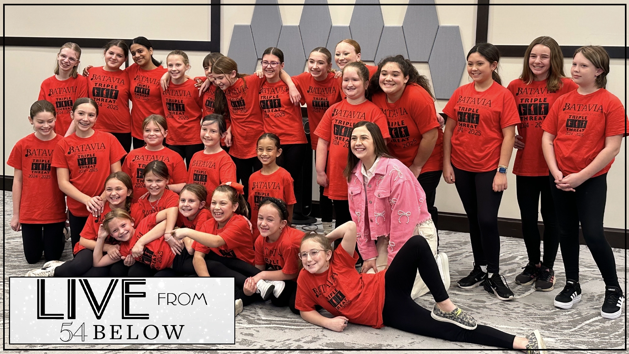 A group of smiling children, mostly girls, pose indoors in matching red 