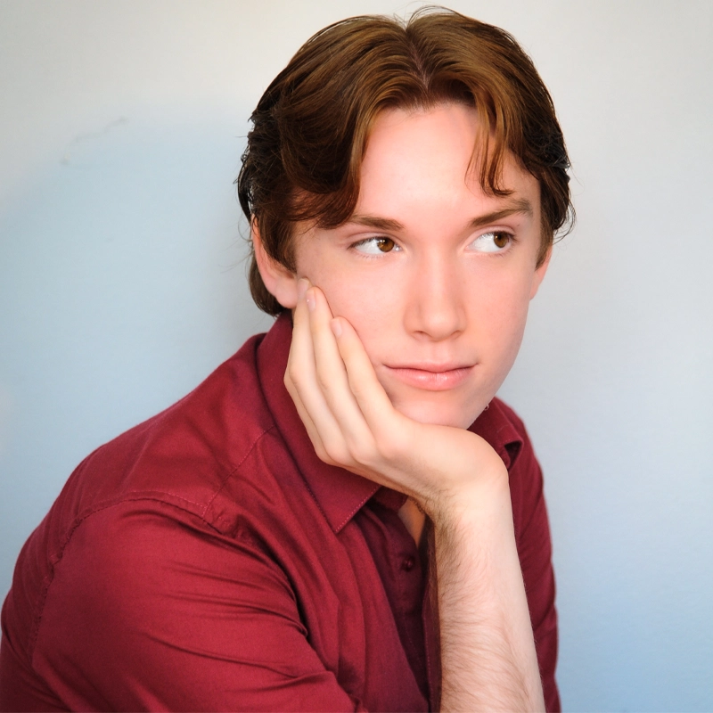 A young person with light skin, medium-length brown hair, and wearing a maroon shirt rests their face on their left hand, gazing thoughtfully to the side against a plain, light-colored background.