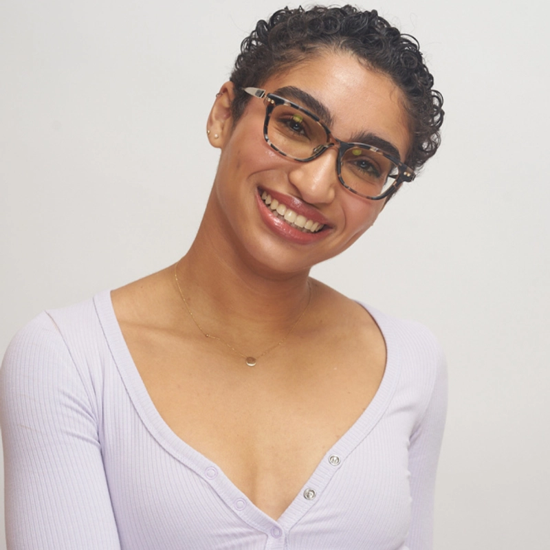 Sumner Lewis, a woman with short curly hair and glasses, smiles at the camera. She wears a light lavender ribbed top with buttons and a delicate gold necklace. The plain white background gives the portrait a bright and clean appearance.