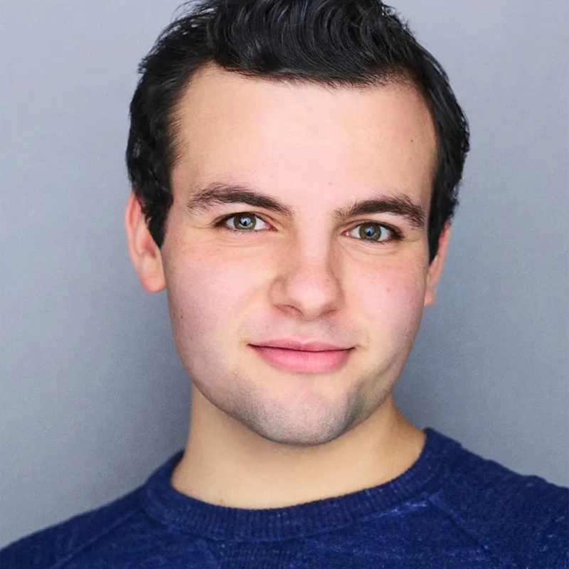 A young man with short dark hair and fair skin looks at the camera, smiling softly. He has blue-green eyes and wears a blue crew-neck sweater. The background is plain and light gray, and the lighting is bright and even.