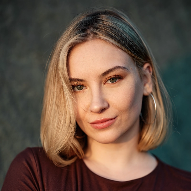 A young woman with straight, shoulder-length blonde hair and hoop earrings looks into the camera with a slight smile. She wears a maroon top and stands against a blurred, dark background, illuminated by soft, natural light.