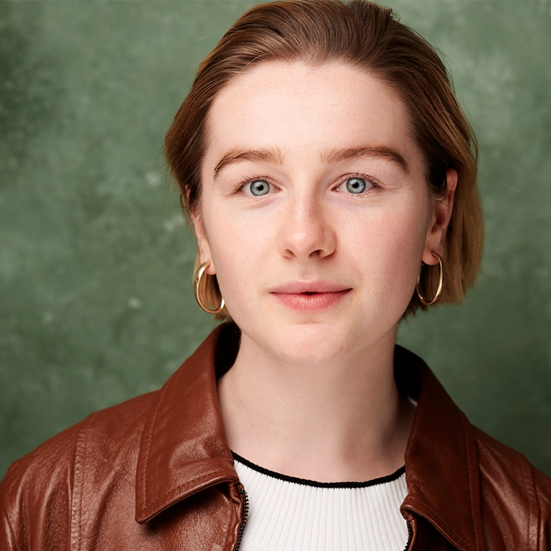 A young person with short, light brown hair and fair skin wears gold hoop earrings, a brown leather jacket, and a white top. They have blue eyes and a slight smile. The backdrop is mottled green; the lighting soft and even—Liz Gilmartin.