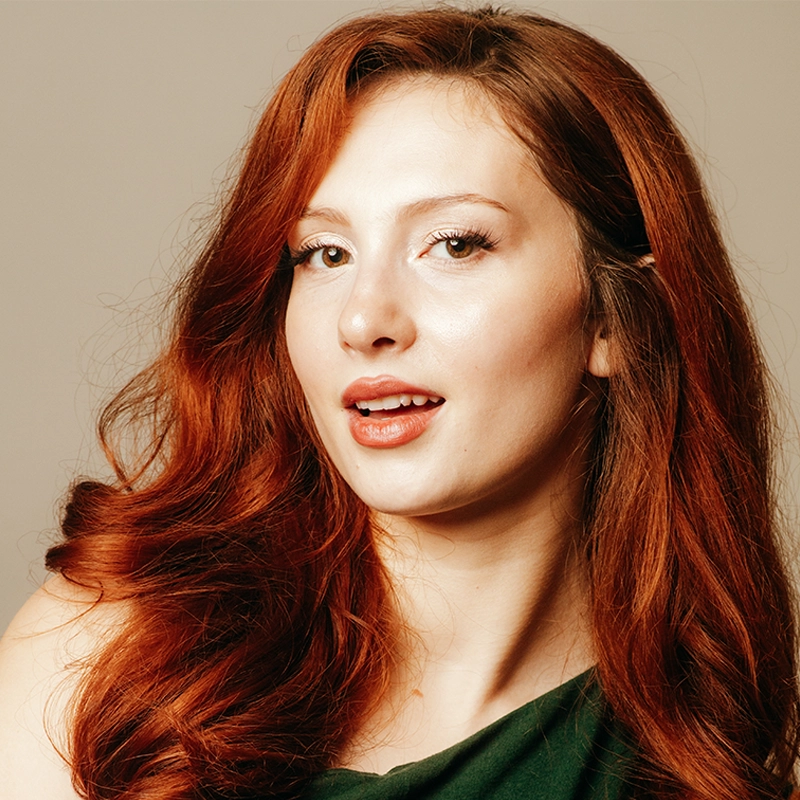 A young woman with long, wavy red hair looks at the camera with a slight smile. She wears a green off-shoulder top and light makeup. The background is plain beige, highlighting her hair and expressive facial features.