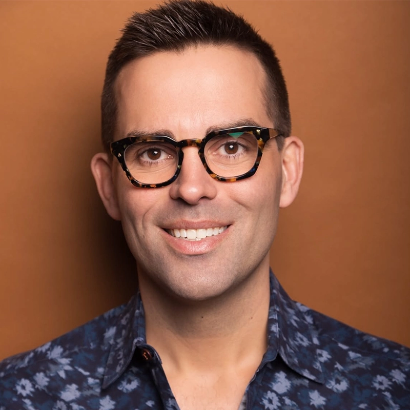 A smiling man with short dark hair and tortoiseshell glasses stands against a brown background. He wears a blue patterned button-up shirt. The soft lighting highlights Luke Frazier as he looks directly at the camera.