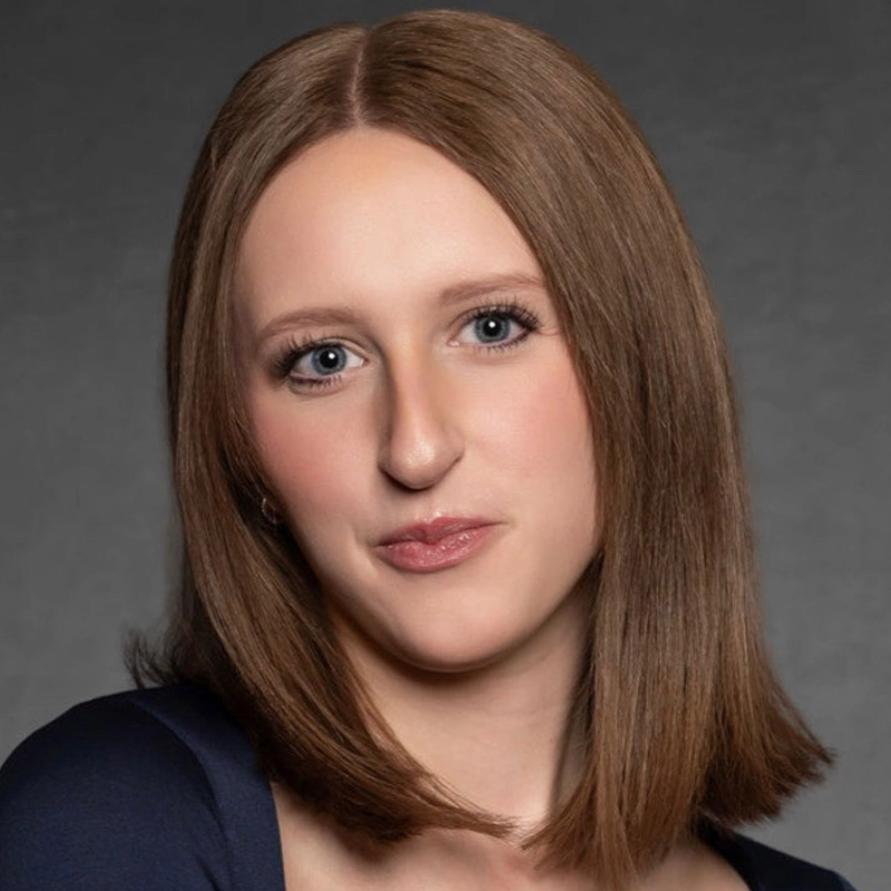 A woman with straight, shoulder-length brown hair and fair skin faces the camera, wearing a navy blue top. She has blue eyes, subtle makeup, and a slight smile. The background is plain and gray, with soft lighting highlighting her features.