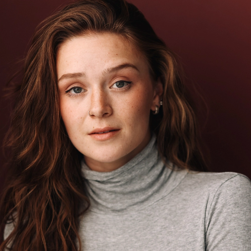 A young woman with long wavy brown hair and fair skin gazes softly at the camera. She wears a light gray turtleneck and small earrings. The background is a warm reddish-brown, creating a calm, neutral portrait setting.