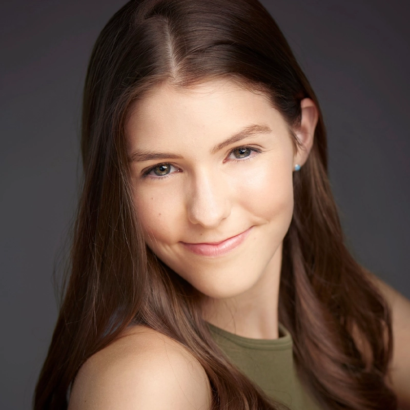 A young woman with long, straight brown hair smiles gently at the camera. She has fair skin, light-colored eyes, and is wearing a sleeveless olive green top. The background is softly blurred in shades of gray, creating a professional portrait look.