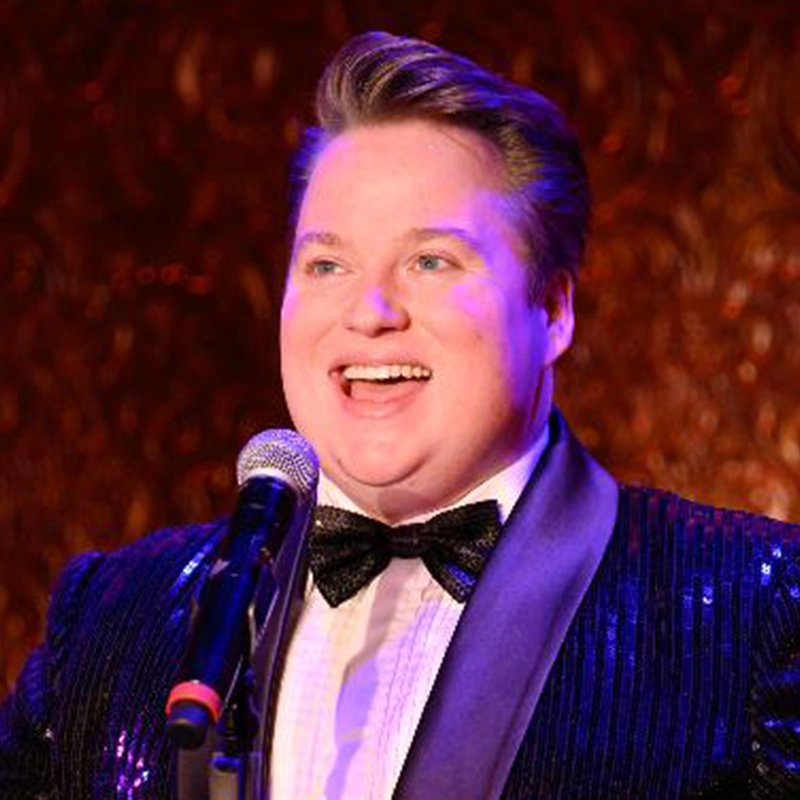 A person with short, styled hair smiles while singing into a microphone. Dressed in a black sequin jacket, black bow tie, and white shirt, Edmund Bagnell performs against a warmly lit, textured reddish-brown background.