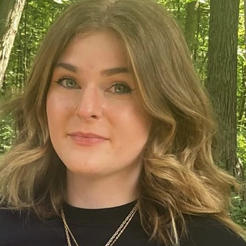 Josie Madzik, a young woman with wavy, light brown hair and green eyes, stands outdoors in a sunlit forest. She wears a black top and layered gold necklaces, smiling slightly as trees with green leaves blur in the background.