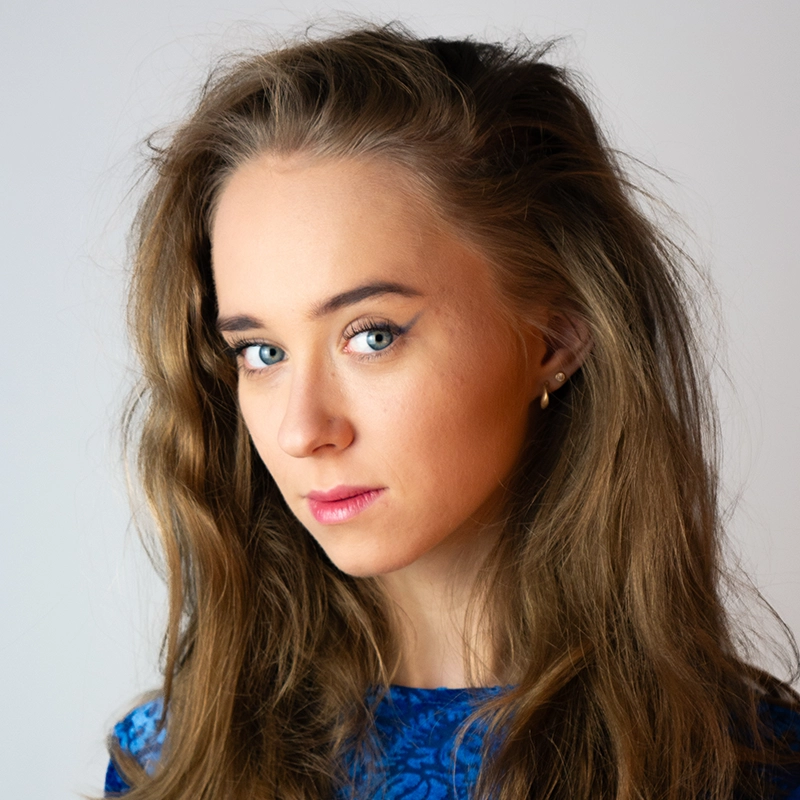 A young woman with long, wavy brown hair, light skin, and blue eyes gazes at the camera. She wears a blue patterned top and small gold hoop earrings. Her makeup includes winged eyeliner and pink lipstick. The background is plain and light-colored.