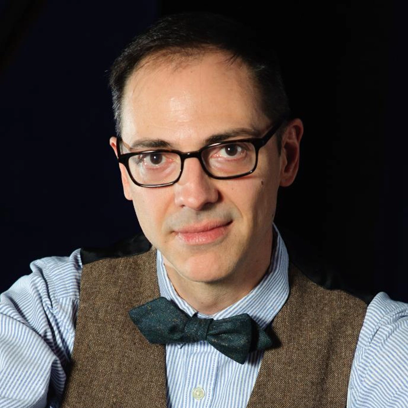 A man with short dark hair and glasses, resembling Edmund Bagnell, wears a brown vest over a blue striped shirt and a dark bow tie, looking directly at the camera against a black background. He has a slight, neutral expression.