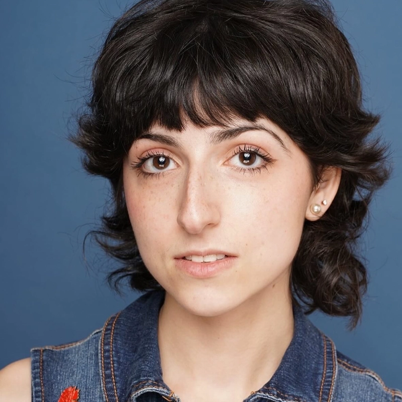 A young person with short, dark, wavy hair and fair skin looks at the camera with a neutral expression. They wear a sleeveless denim top and small stud earrings. The background is a solid blue color.