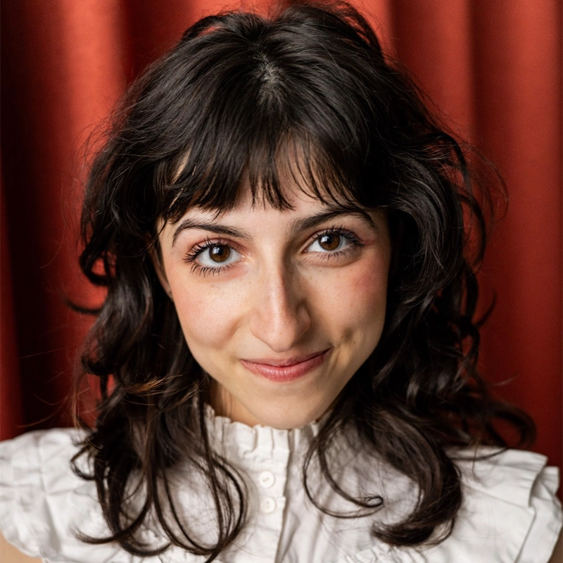 A person with wavy dark hair and bangs smiles softly at the camera. The person is wearing a white ruffled blouse and stands in front of a red curtain, with warm lighting.