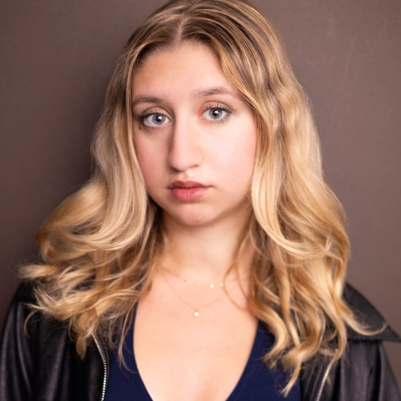 A young woman, Kailyn Marra, with wavy blonde hair and light eyes stands in front of a brown background. She wears a black jacket over a dark top and delicate gold necklaces, her expression neutral as she looks directly at the camera.