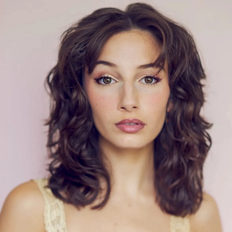 Julia Martin, a young woman with wavy, medium-length brown hair and hazel eyes, looks directly at the camera. She wears natural makeup with soft pink lipstick and gold eyeshadow, a light lacy top, and stands against a pale, neutral background.