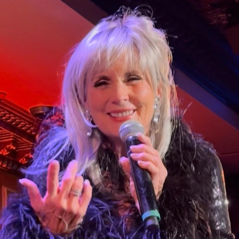 An older woman with long silver hair smiles warmly, holding a microphone in one hand and gesturing with the other. Channeling her inner Edmund Bagnell, she wears sparkling earrings and a black feathered outfit under warm red and purple stage lights.