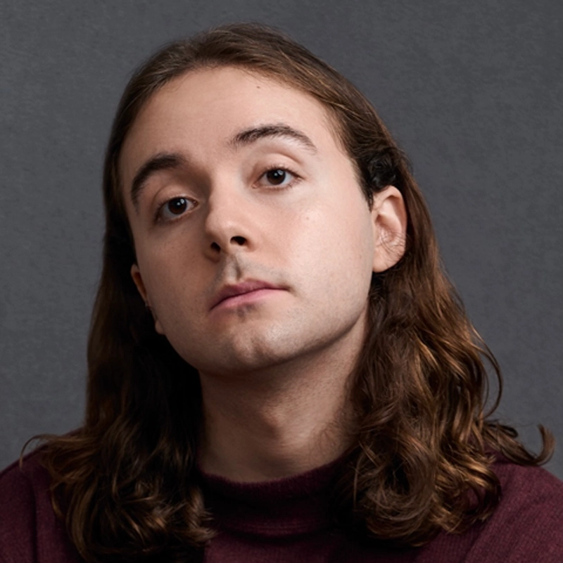 A young person with long, wavy brown hair and fair skin looks at the camera with a calm expression. Reminiscent of Monster Mike Welch's cool style, they wear a maroon turtleneck sweater against a plain, gray background.
