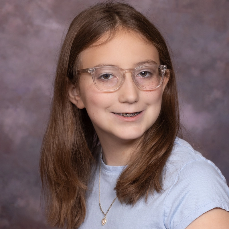 Adalinn McQueen, a young girl with long brown hair, wears clear glasses, a light blue shirt, and a necklace with a small pendant. She smiles brightly, showing braces, posed in front of a mottled purple and gray studio background.