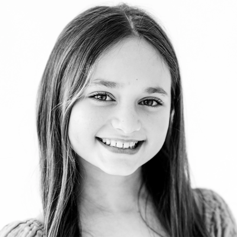 Black and white portrait of young Maggie Meisel with long straight hair, smiling softly at the camera. She has visible teeth, expressive eyes, and wears a textured top. The plain white background emphasizes her face and natural expression.