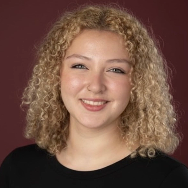 Dominique Alvarez, a young woman with light skin and curly, shoulder-length blonde hair, smiles at the camera. She wears a black top and a small nose ring. The background is a solid maroon color.