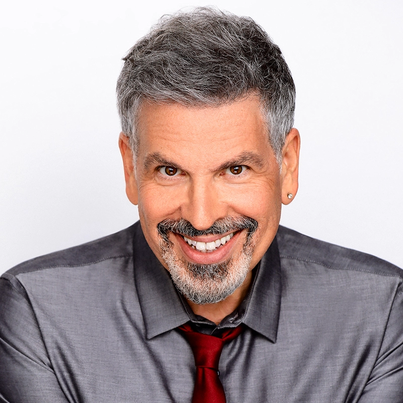 A middle-aged man with short gray hair, a goatee, and mustache smiles warmly at the camera. He wears a metallic gray dress shirt with a red tie and a small hoop earring in his left ear—reminiscent of Edmund Bagnell. The background is plain white.