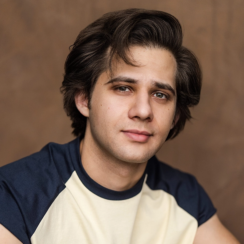 A young man, Michael Raul Perez, with medium-length brown hair and a slight smile poses against a plain brown background. He is wearing a navy blue and cream raglan t-shirt and looking directly at the camera with a relaxed expression.