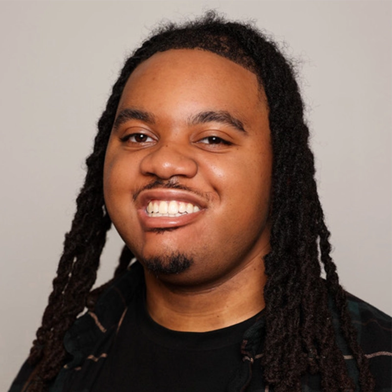 A smiling person with long, dark dreadlocks and a short beard faces the camera. Channeling the cool style of Monster Mike Welch, they wear a dark shirt and plaid outerwear against a plain light gray studio background.