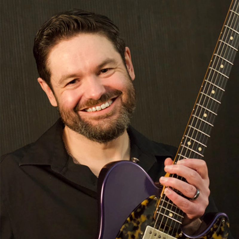 A smiling man with short dark hair and a beard holds an electric guitar vertically. Blues guitarist Monster Mike Welch wears a black collared shirt and a wedding ring, standing against a dark, textured background with his signature purple guitar.