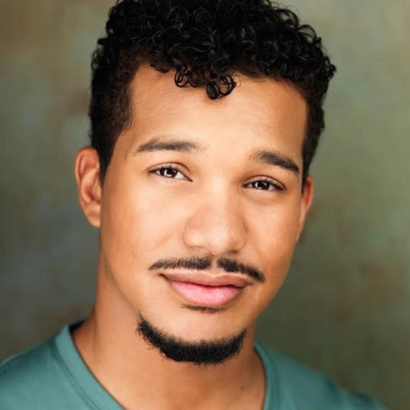 Da’Von T. Moody, a young man with short, curly dark hair and a trimmed goatee, smiles softly. He has warm brown skin, expressive eyes, and wears a teal shirt against a softly blurred green and brown background for a natural feel.