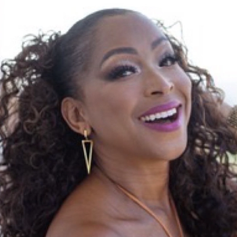 A woman with long, curly brown hair smiles broadly. She wears bold purple lipstick, dramatic eye makeup, gold geometric earrings, and a gold necklace. The background is softly blurred, highlighting her cheerful expression and glamorous style.