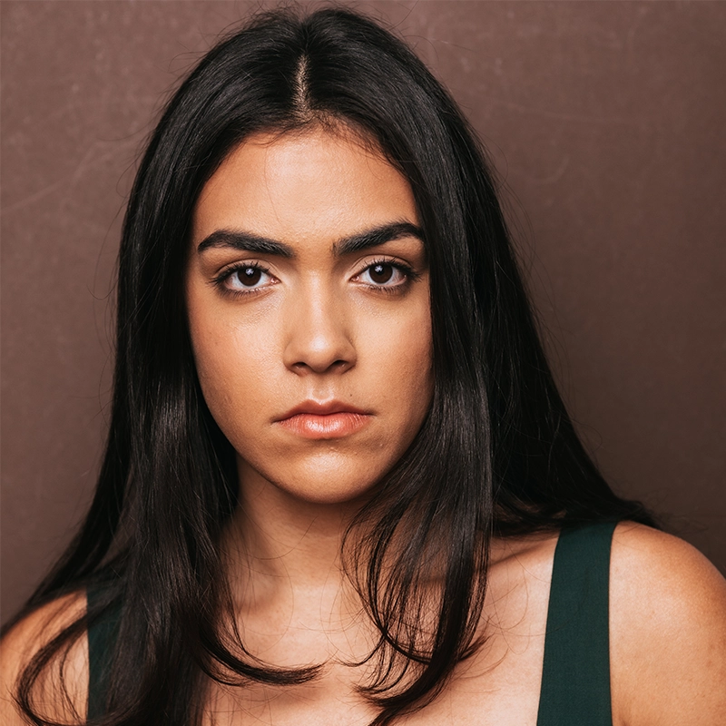 A young woman with long, straight dark hair and brown eyes stares directly at the camera with a serious expression. She wears a dark green sleeveless top and stands against a plain brown background. The lighting is soft and even.