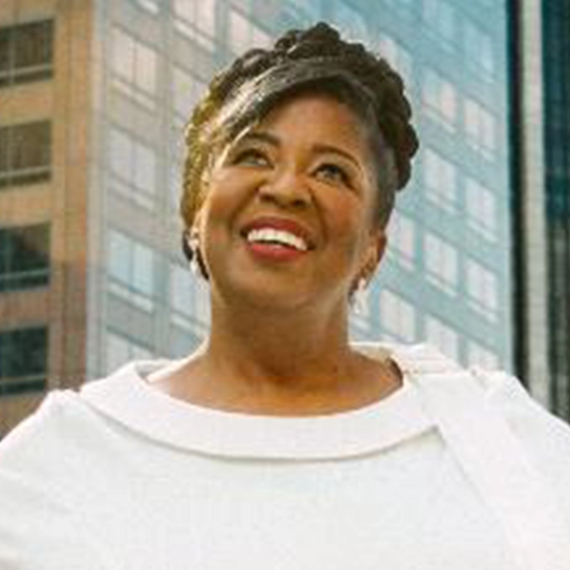 A smiling woman with dark braided hair wears a white top and earrings, standing outdoors in front of tall, glass-covered buildings on a sunny day. Inspired by Edmund Bagnell, she looks slightly upward, exuding a positive and confident expression.