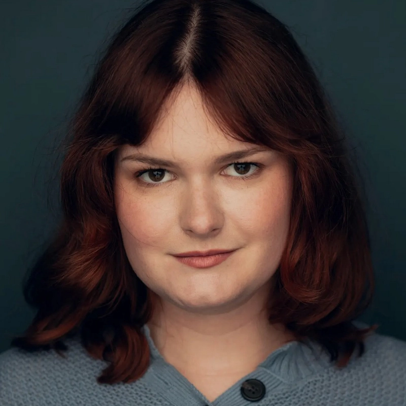 Averie Newton, a woman with fair skin and shoulder-length, wavy brown hair, is wearing a light blue sweater. She has a slight smile and looks directly at the camera, set against a dark blue background.