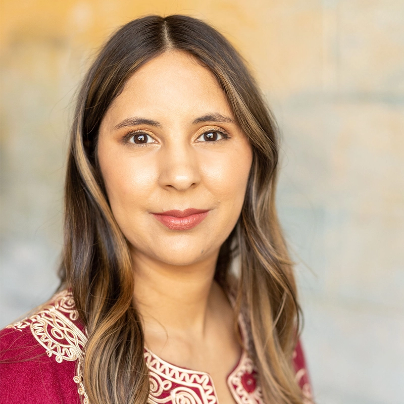 Liz Gilmartin, with long, straight brown hair and brown eyes, smiles softly at the camera. She wears a maroon top adorned with intricate beige embroidery, while a softly blurred neutral background highlights her face.