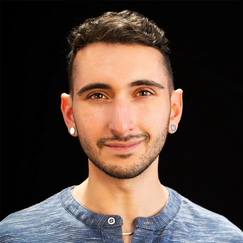 A young person with short, curly dark hair and trimmed facial hair smiles gently. Alex Mogil Cooke wears round earrings, a blue henley shirt, and stands against a solid black background with soft lighting on their face.