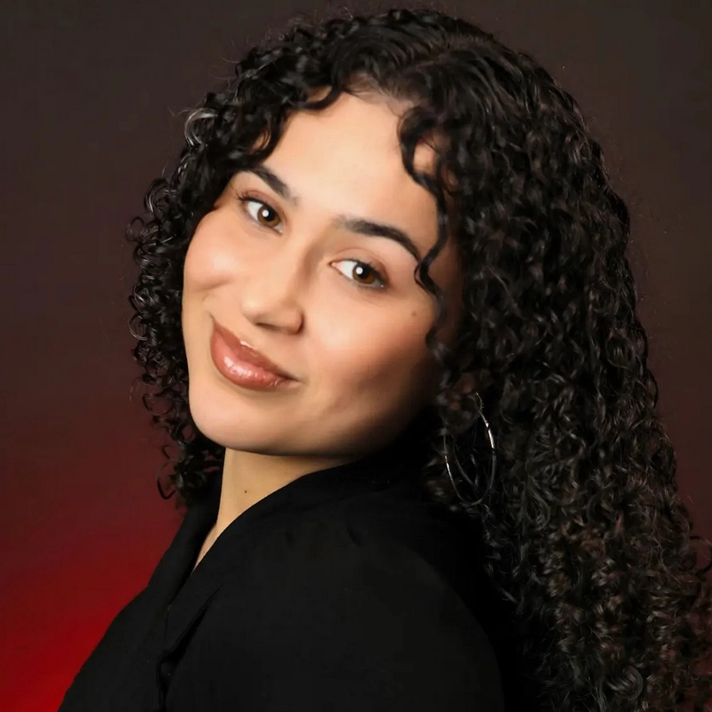 Moira O'Connor, with medium skin and dark curly hair, smiles gently as she glances over her shoulder. She wears a black top and large hoop earrings, set against a gradient of dark red to black for a warm, elegant atmosphere.