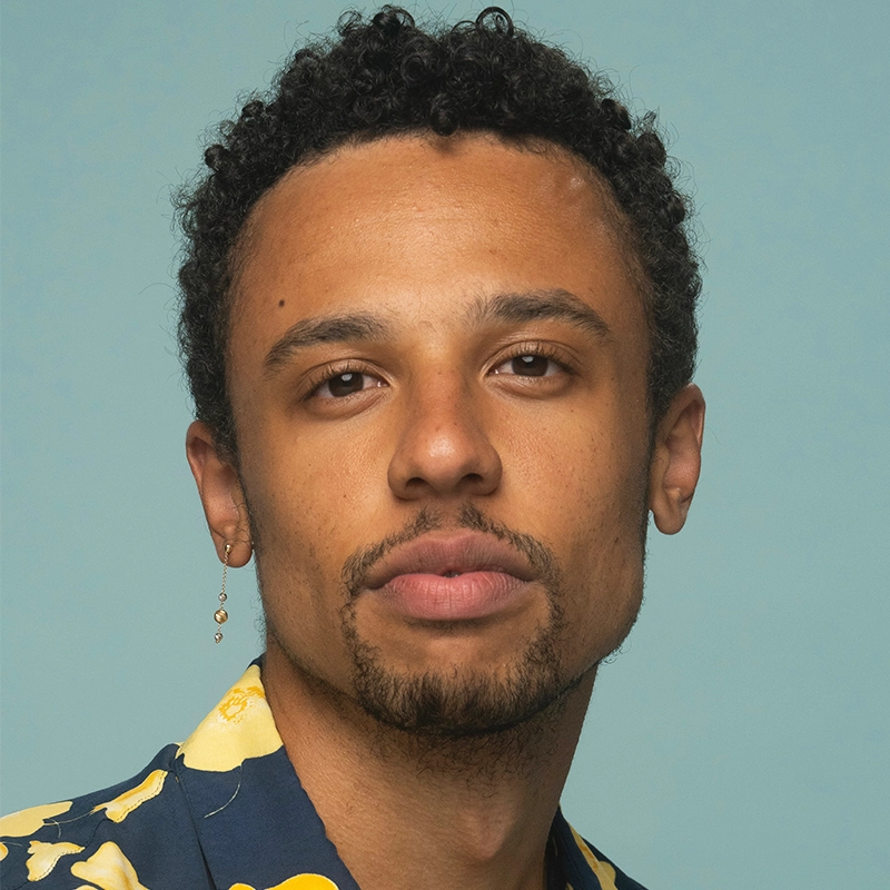 A person with short curly hair and light brown skin looks directly at the camera. They have a short beard, mustache, and are wearing a dangling earring in their left ear. Taylor Leccese wears a dark shirt with a yellow floral pattern against a light blue background.