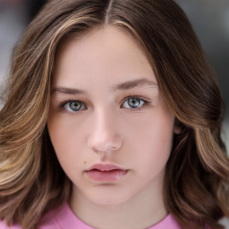 A young person with wavy, light brown hair and blue eyes looks directly at the camera. They have clear skin, natural makeup, and are wearing a light pink top. The background is softly blurred in neutral tones.