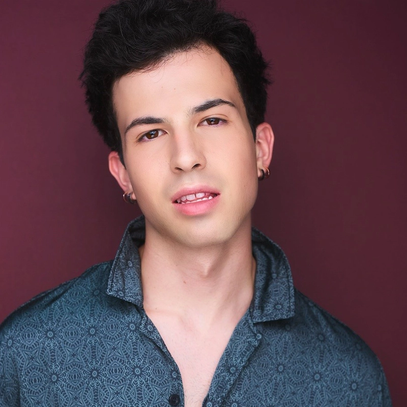 A young man, Ryan Orbe-Basch, with short, curly dark hair poses against a burgundy background. He wears a patterned blue shirt with the collar slightly open and small hoop earrings, facing forward with a relaxed, neutral expression.
