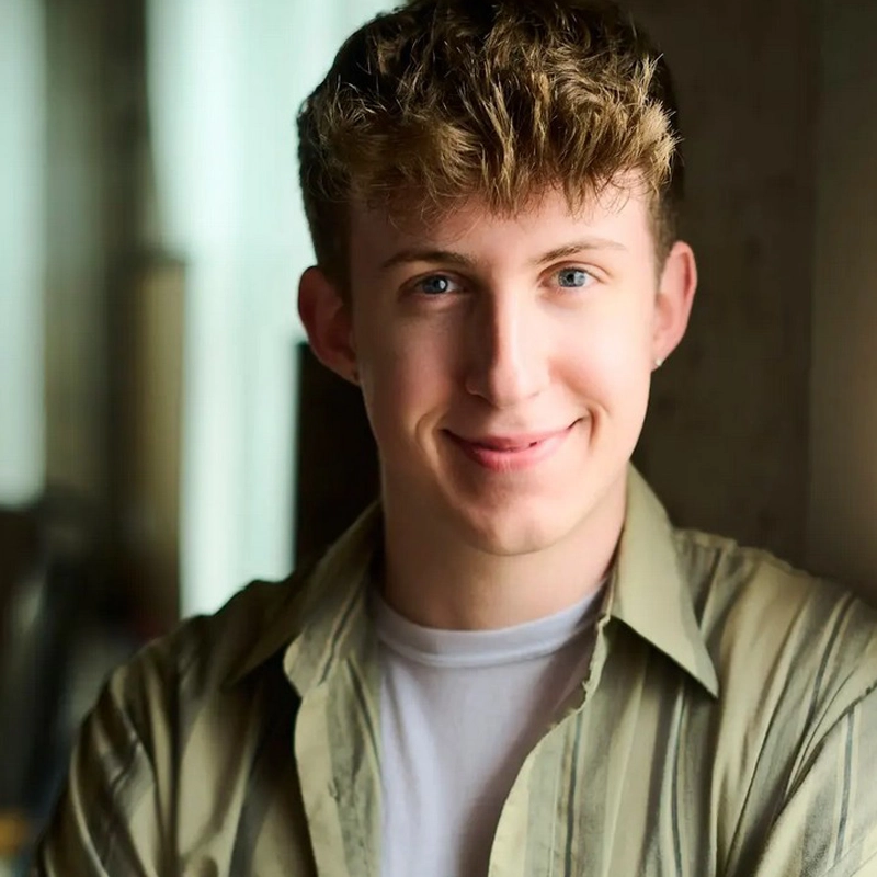A young person with short, curly blond hair and blue eyes smiles warmly. They wear a light green striped shirt over a white t-shirt and stand in soft, natural light with a blurred indoor background. Their expression is friendly and relaxed.