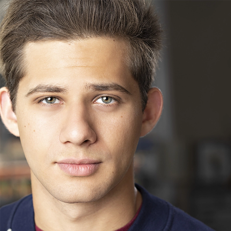 A young man with short, brown hair looks directly at the camera with a neutral expression. He has light skin, hazel eyes, and wears a dark shirt over a red top. The background is softly blurred, drawing focus to his face.