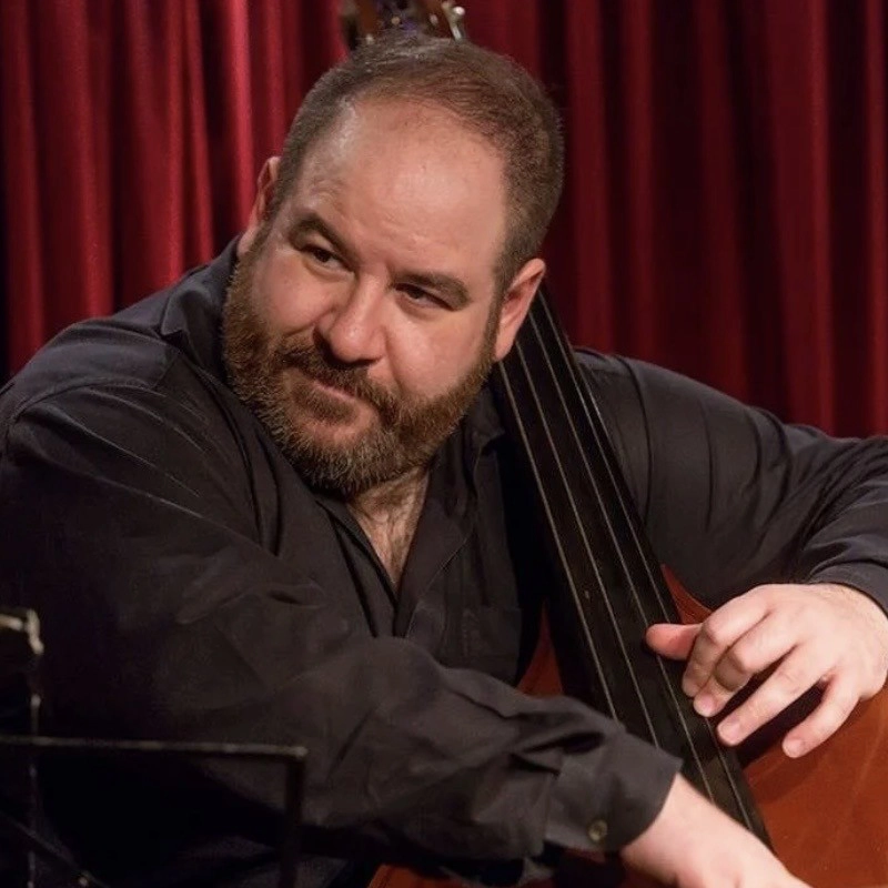 A bearded man in a black shirt plays a double bass on stage. He smiles slightly and looks to his left. Behind Marco Panascia, deep red stage curtains provide a dramatic backdrop, creating a warm and intimate performance atmosphere.