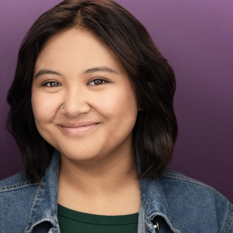 Isabella Paragas, with medium-length dark hair, smiles gently in front of a purple background. She wears a denim jacket over a green top and has a nose piercing. Soft lighting highlights her clear skin and friendly expression.