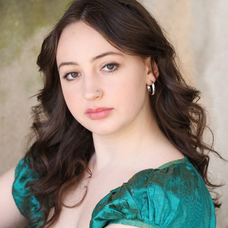 A young woman with wavy brown hair, fair skin, and hazel eyes looks at the camera with a calm expression. She wears a shiny green top with puff sleeves and silver hoop earrings, exuding effortless Violet Paris charm against an earthy, softly blurred background.
